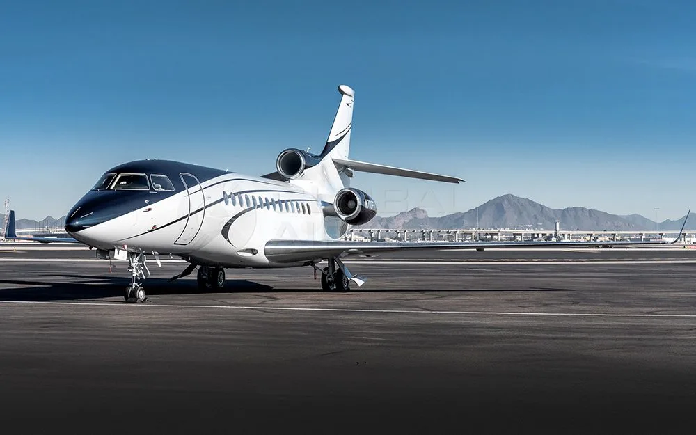 A Dassault Falcon 900EX private jet with a dark blue and white paint scheme parked on an airfield with mountains in the background.