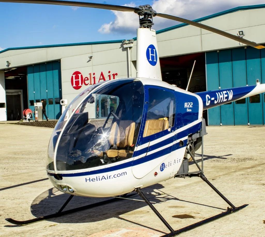 A white and blue Robinson R22 Beta helicopter (G-JHEW) with "Heli Air" branding parked on a tarmac in front of hangars.