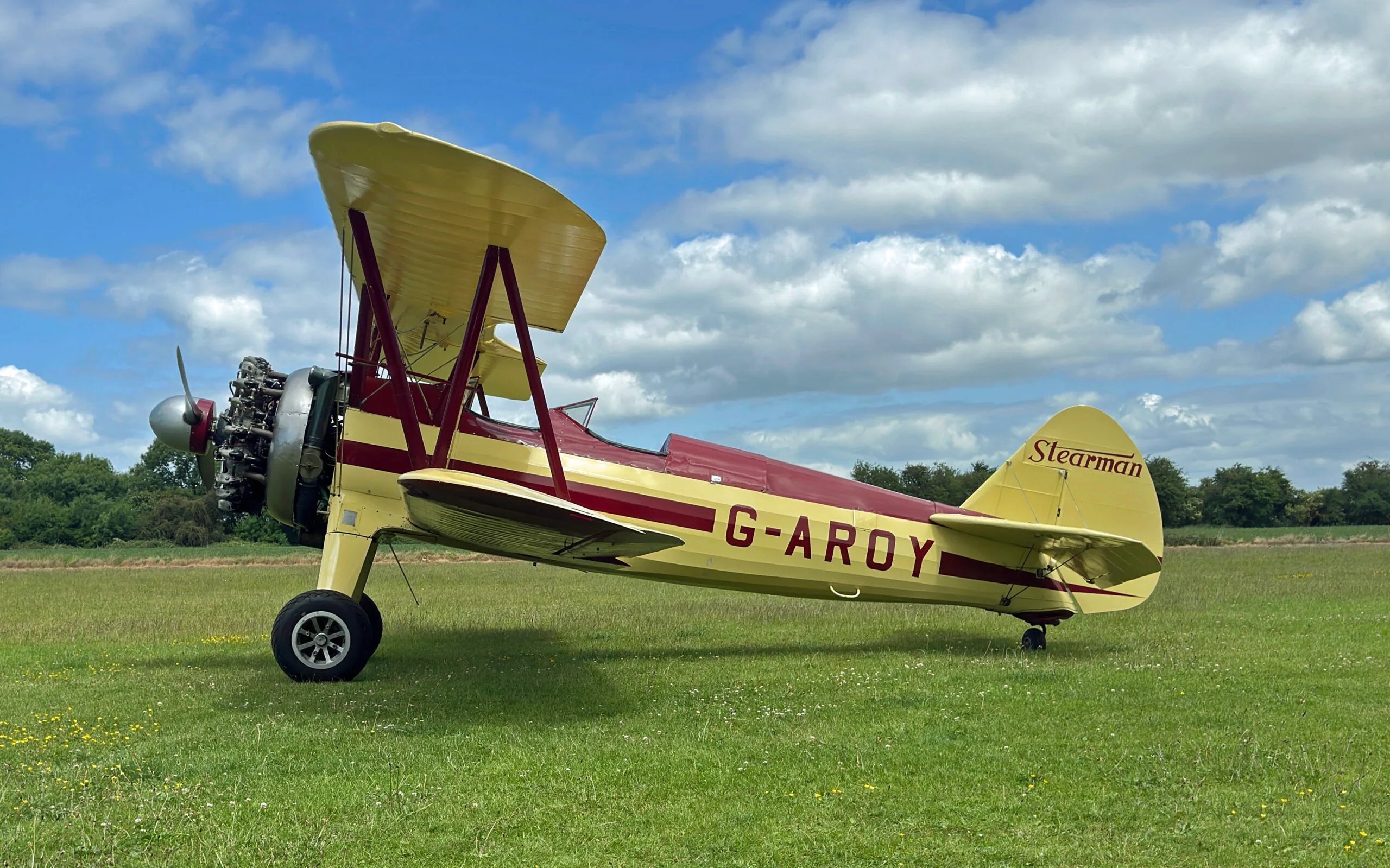 A yellow and red Stearman biplane, G-AROY, seen from the side on a grassy field with clouds in the sky.