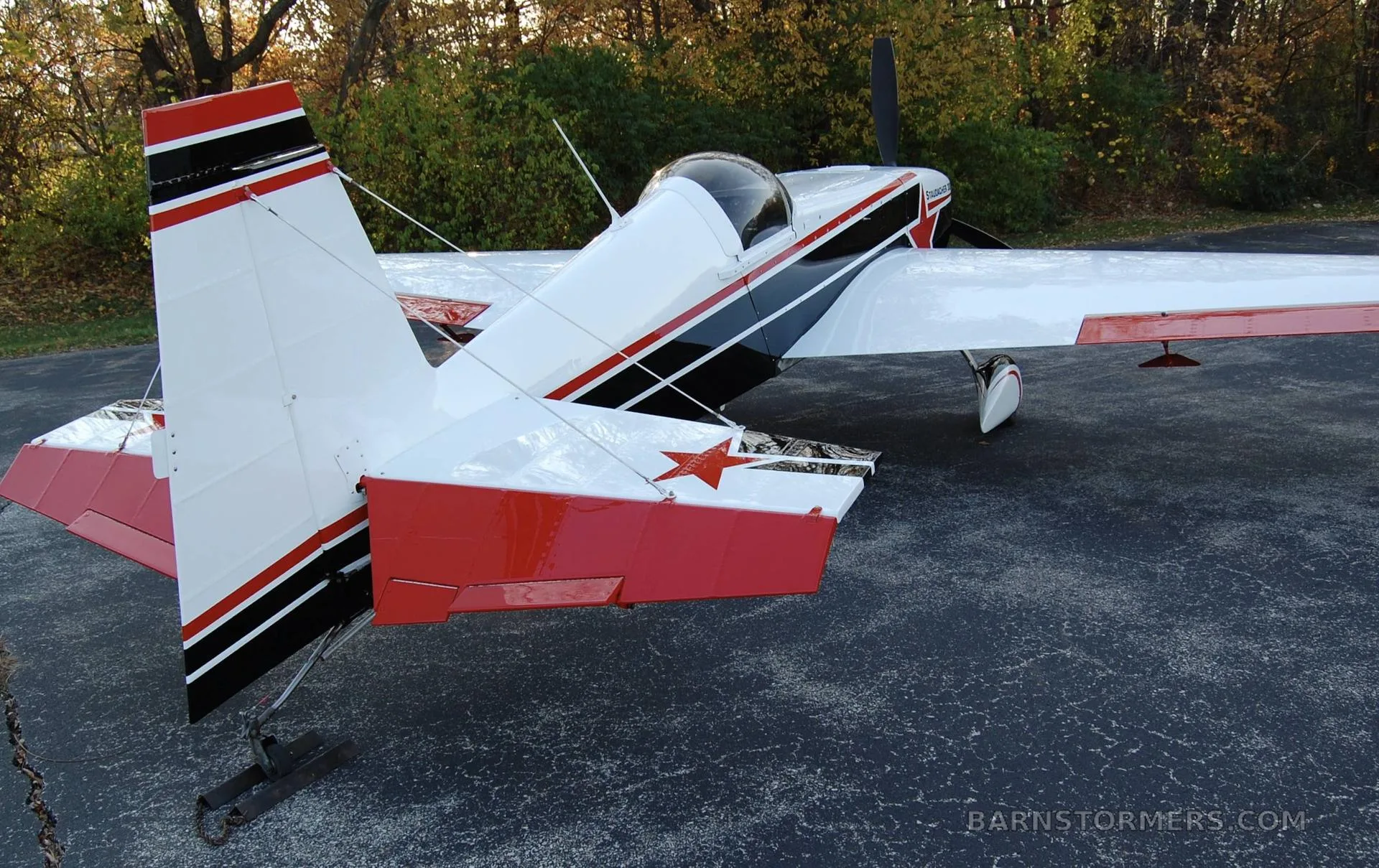 A white, black, and red Staudacher 300 aerobatic aircraft is parked on an asphalt surface with autumn trees in the background, viewed from the rear-right.
