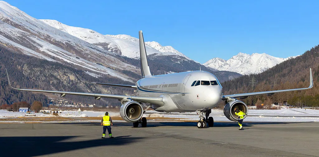 A silver Airbus A320 family aircraft on an airport tarmac with snow-capped mountains in the background, with two ground crew members nearby.