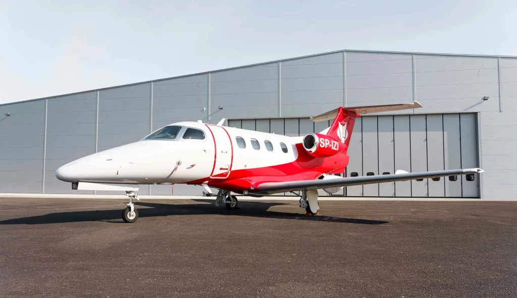 A white and red Embraer Phenom 300, registration SP-IZI, parked on an airport tarmac in front of a large hangar.