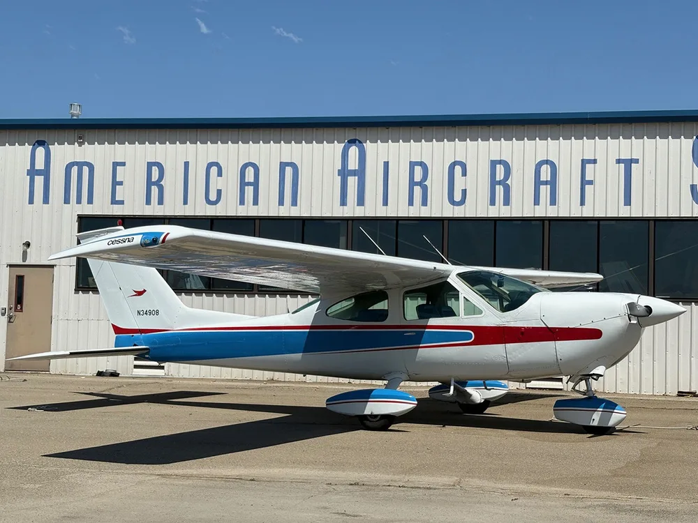 A white Cessna 177 Cardinal with blue and red stripes parked on an asphalt ramp in front of a hangar.