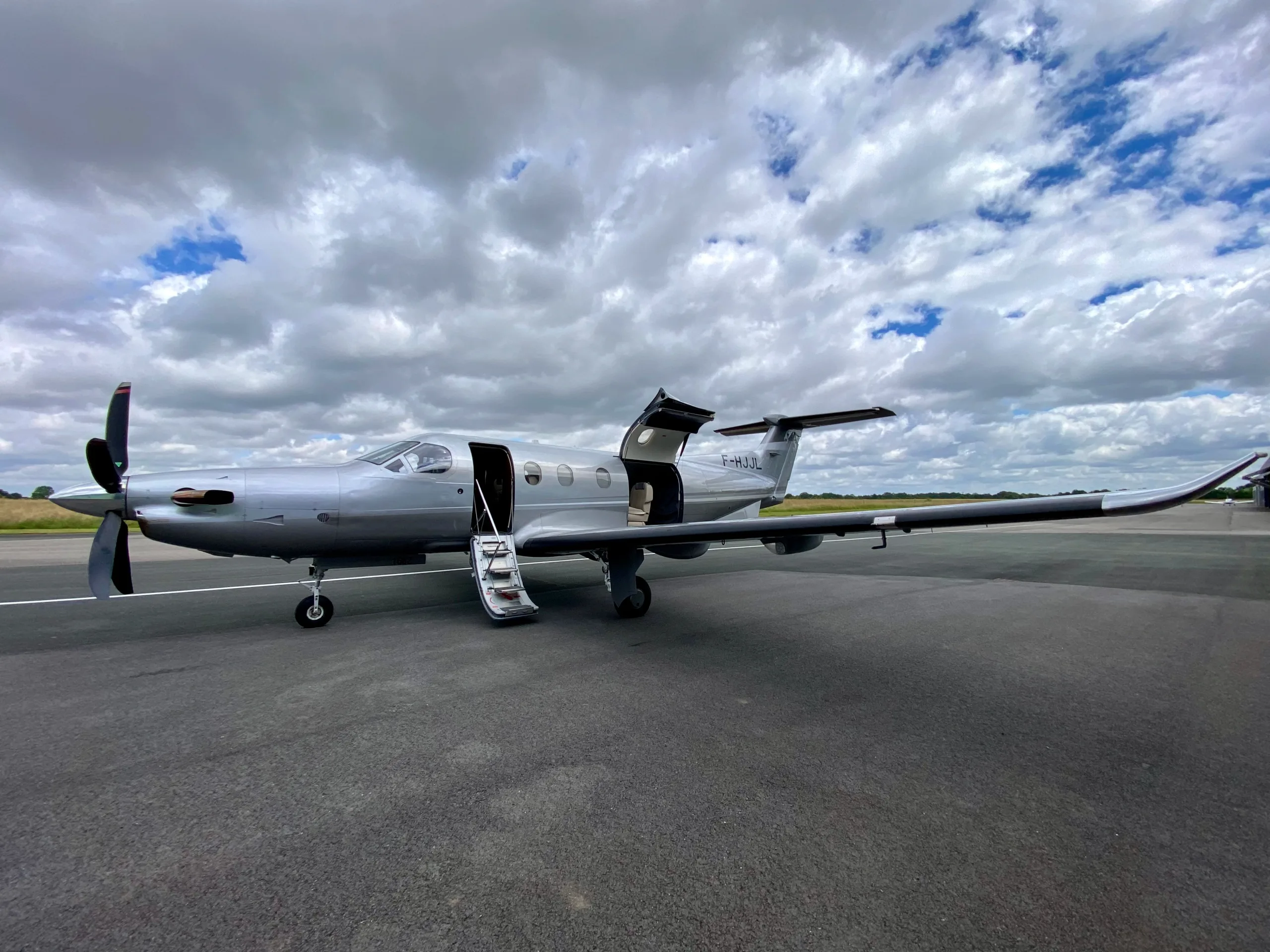 A silver Pilatus PC-12 aircraft with its main and cargo doors open, parked on an asphalt runway under a cloudy sky.