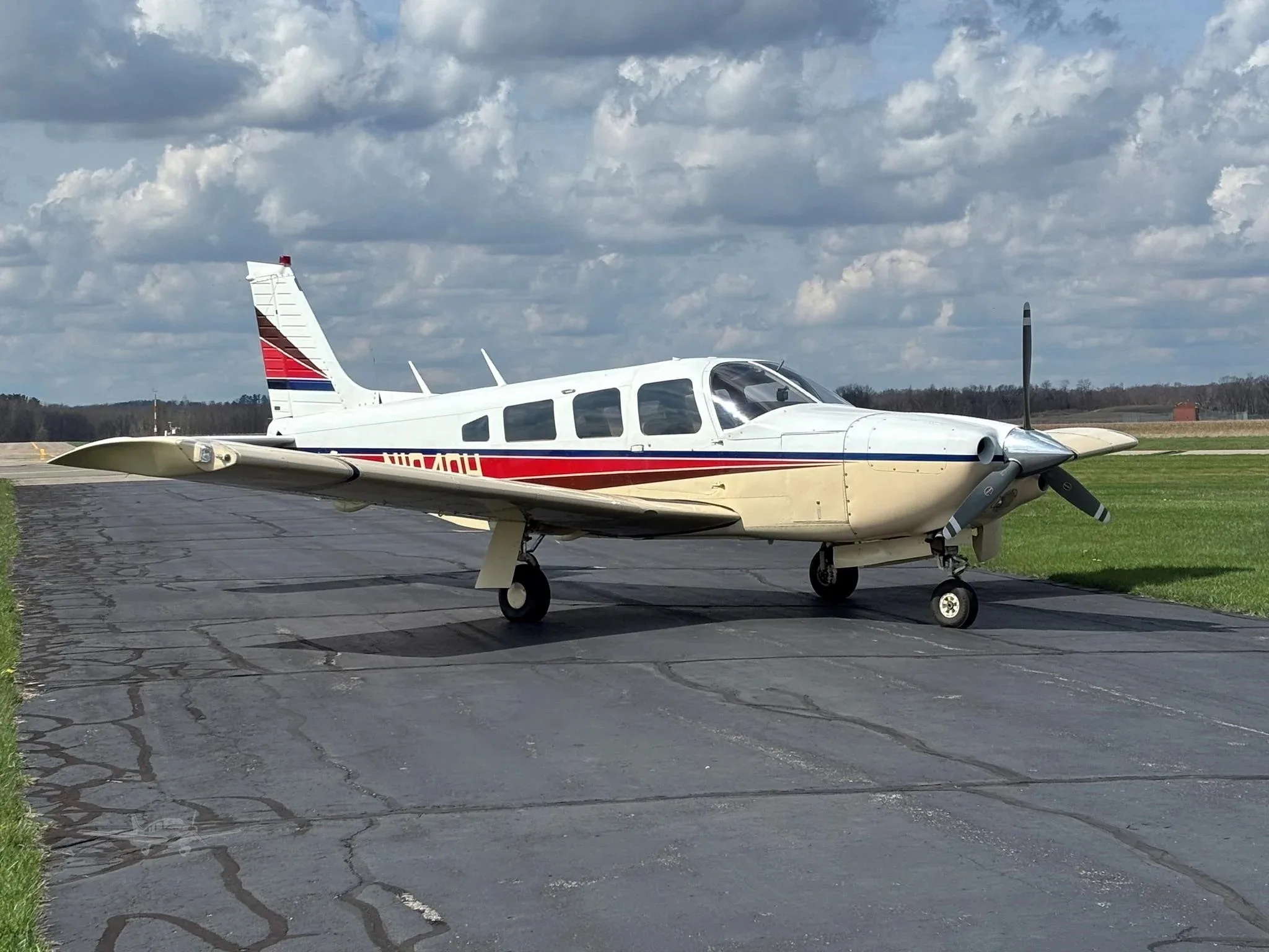 A white and beige single-engine aircraft with red and blue stripes on the tail and fuselage, parked on an asphalt runway under a cloudy sky.
