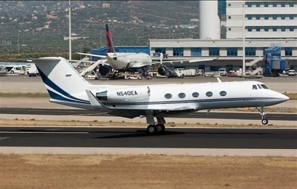 A white Gulfstream G450 business jet with blue stripes is seen on a runway at an airport, with a Delta aircraft in the background.