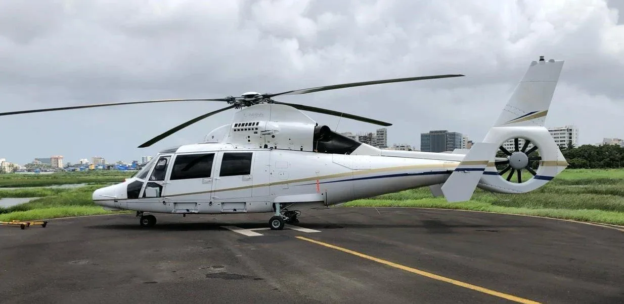 A white helicopter with gold and blue accents parked on a tarmac with buildings and greenery in the background.