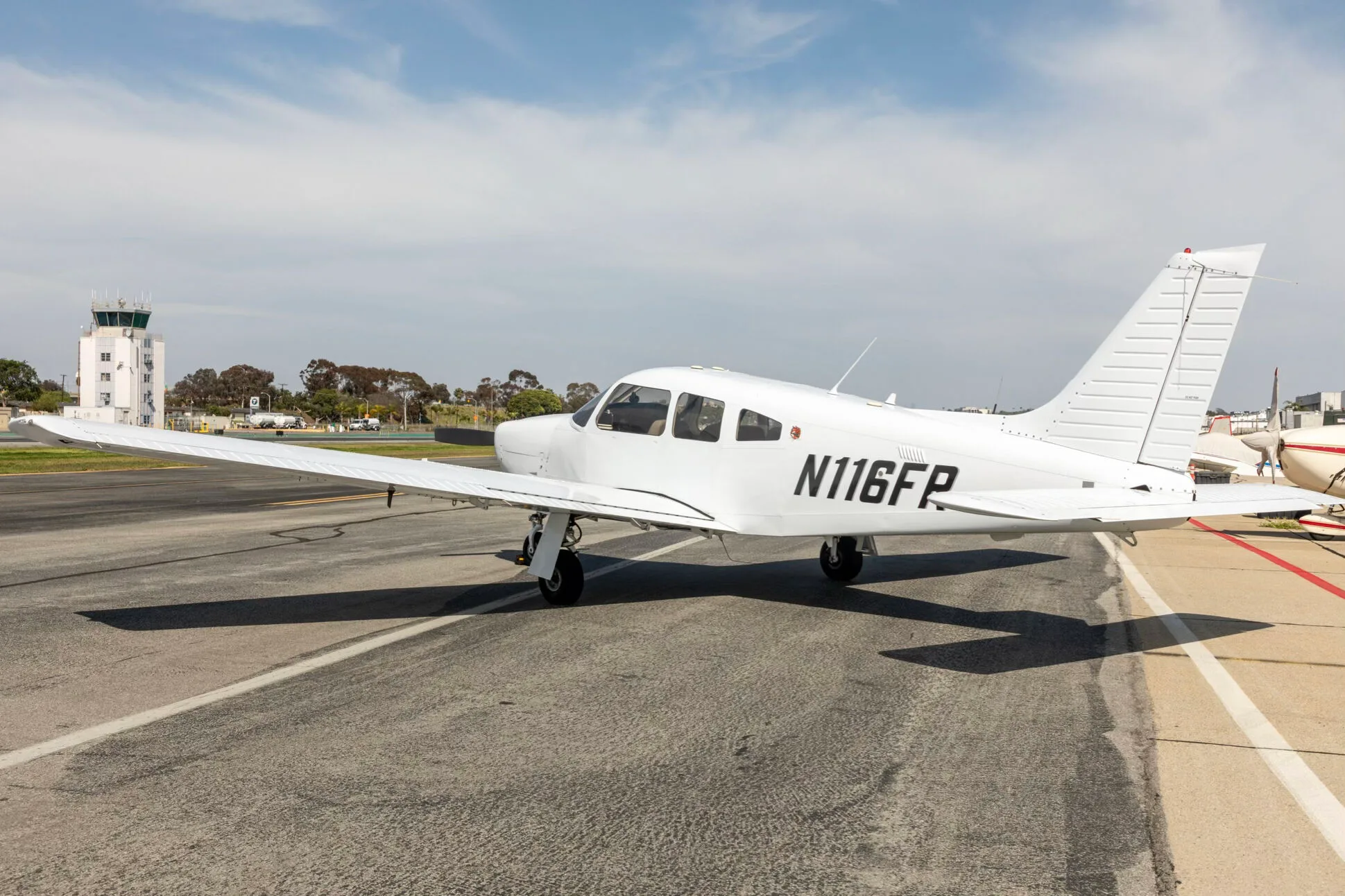 A white Piper PA-28-181 Archer III aircraft, registration N116FR, parked on an airfield with a control tower in the distance.