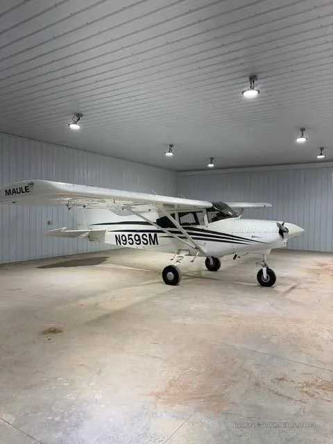 A white Maule M-7-235B aircraft with black stripes and registration N959SM is parked inside a hangar.