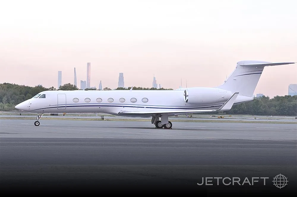 A white Gulfstream G650 business jet with blue stripes on a tarmac, with a city skyline in the background.