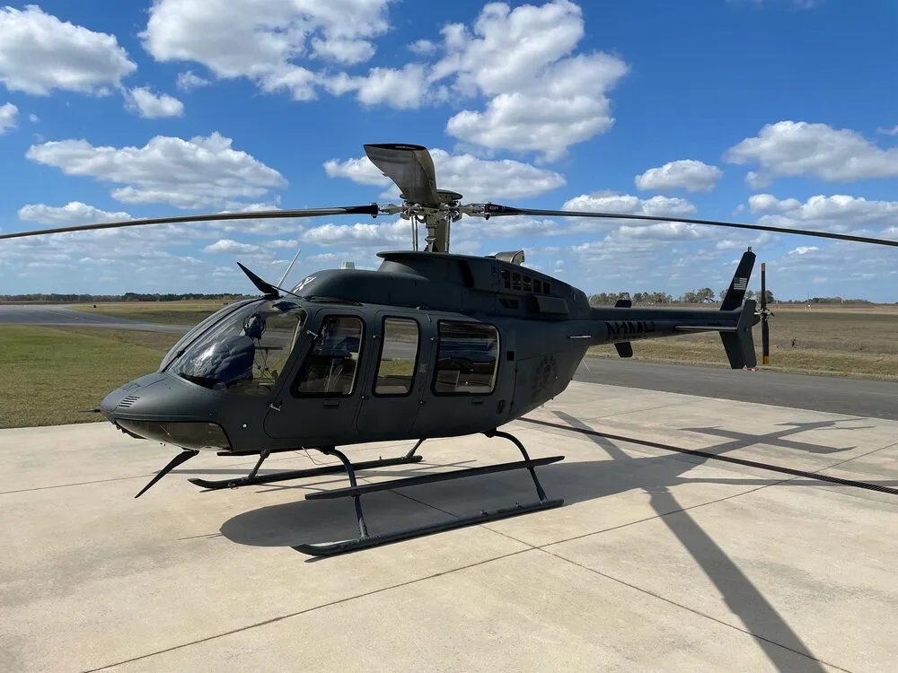 A dark grey Bell 407 helicopter, tail number N1MH, parked on a concrete pad at an airfield under a partly cloudy sky.