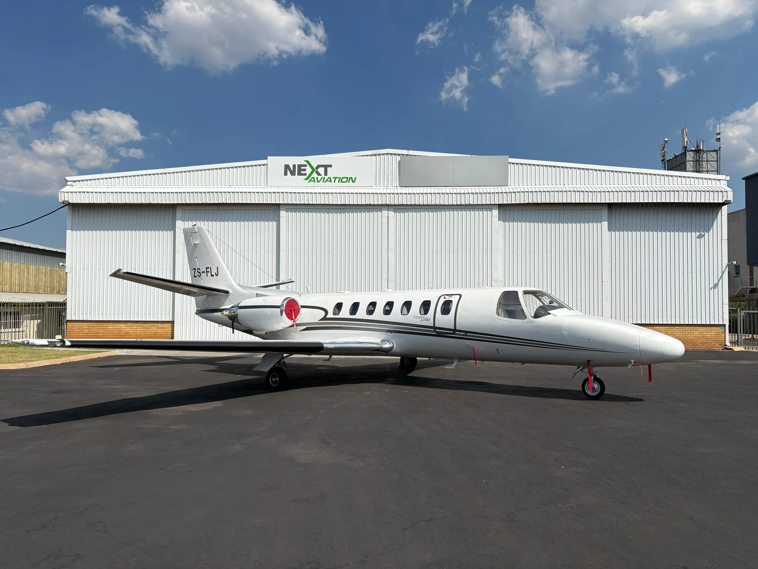 A white Cessna Citation Ultra business jet with black and grey stripes is parked on an asphalt apron in front of a white hangar under a blue sky.