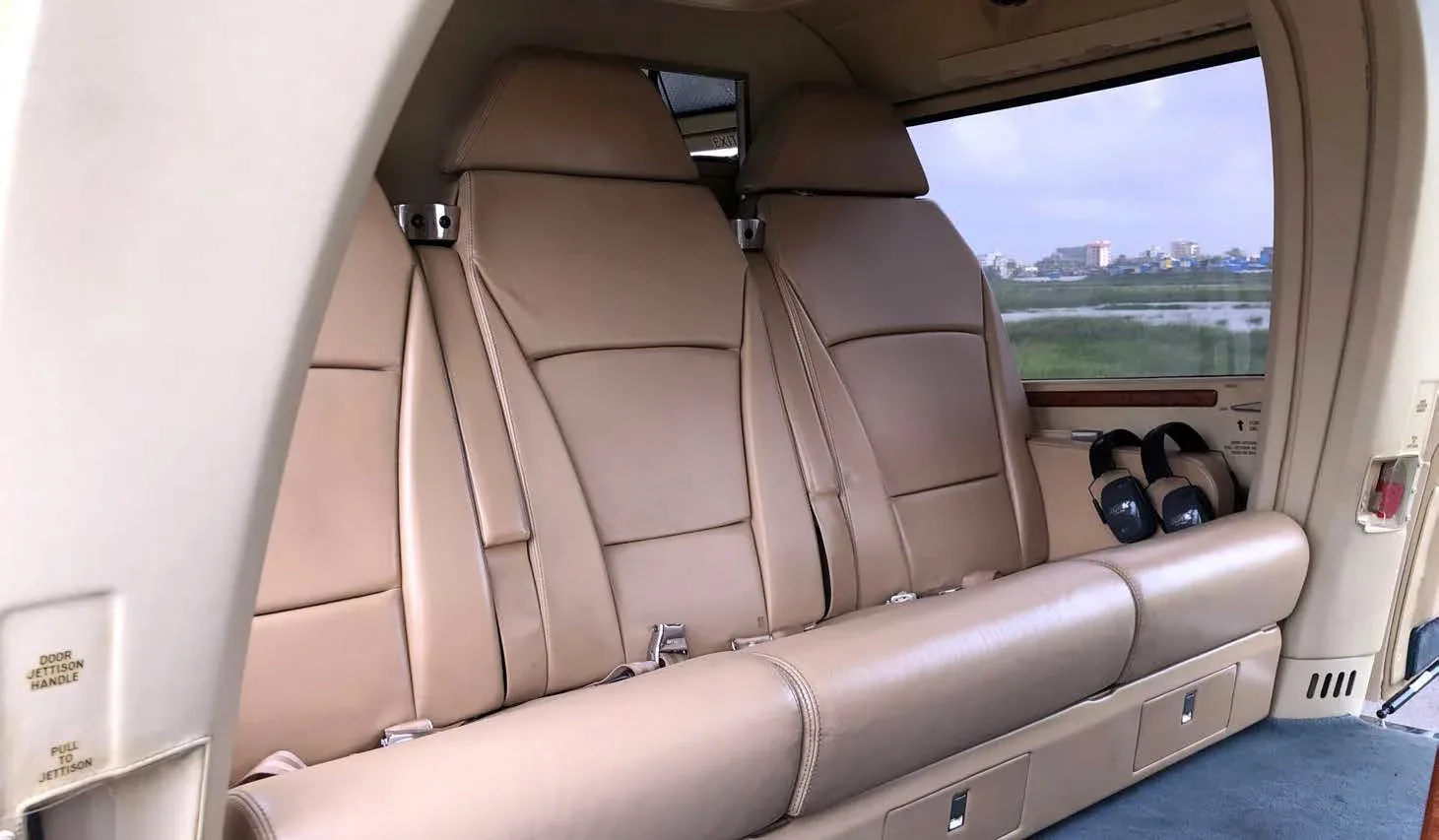 A row of two beige leather passenger seats in a helicopter cabin, with a window view of a distant city and green landscape.