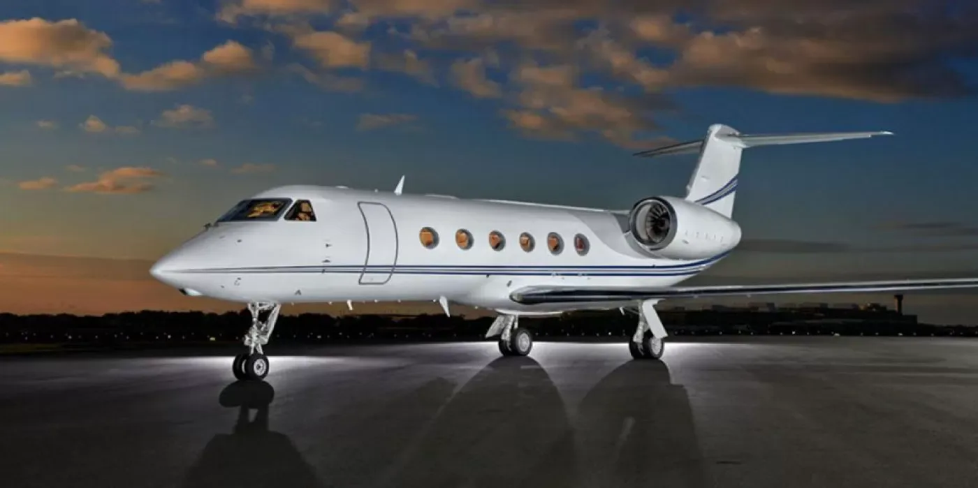 A white Gulfstream-style business jet with blue stripes parked on a runway at sunset or sunrise.