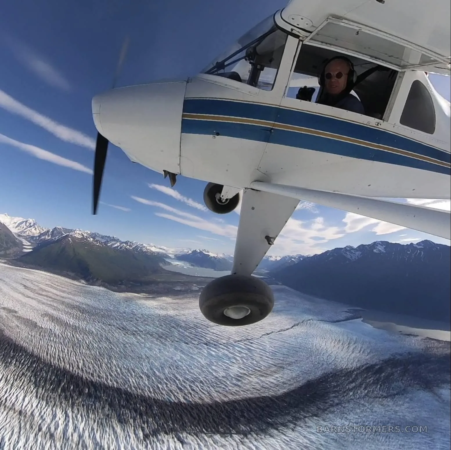 A high-wing aircraft with a pilot visible in the cockpit flying over a vast glacier and mountains under a blue sky.