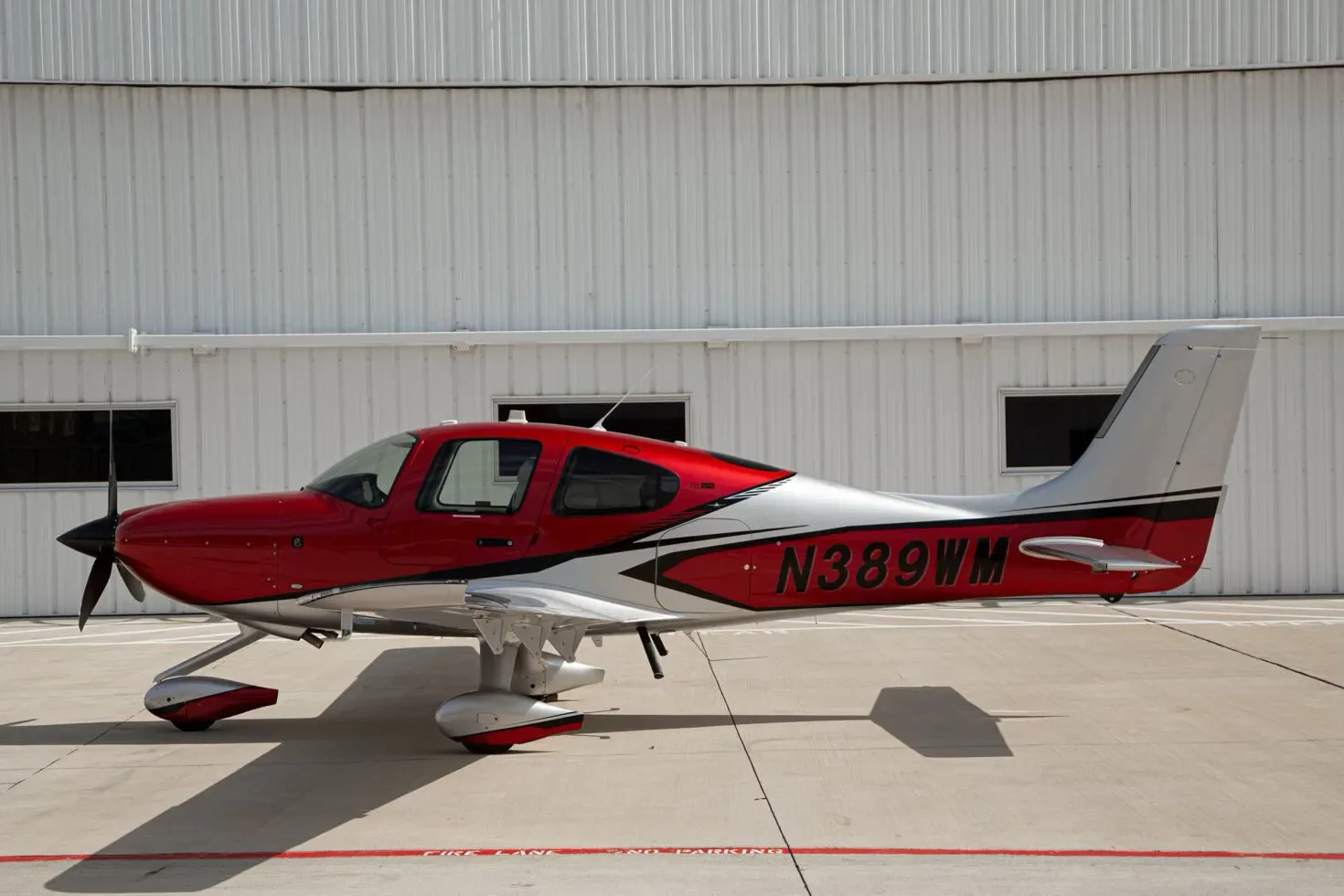 A side view of a red and silver single-engine propeller aircraft, N389WM, with its doors closed, parked on an airfield in front of a white hangar.