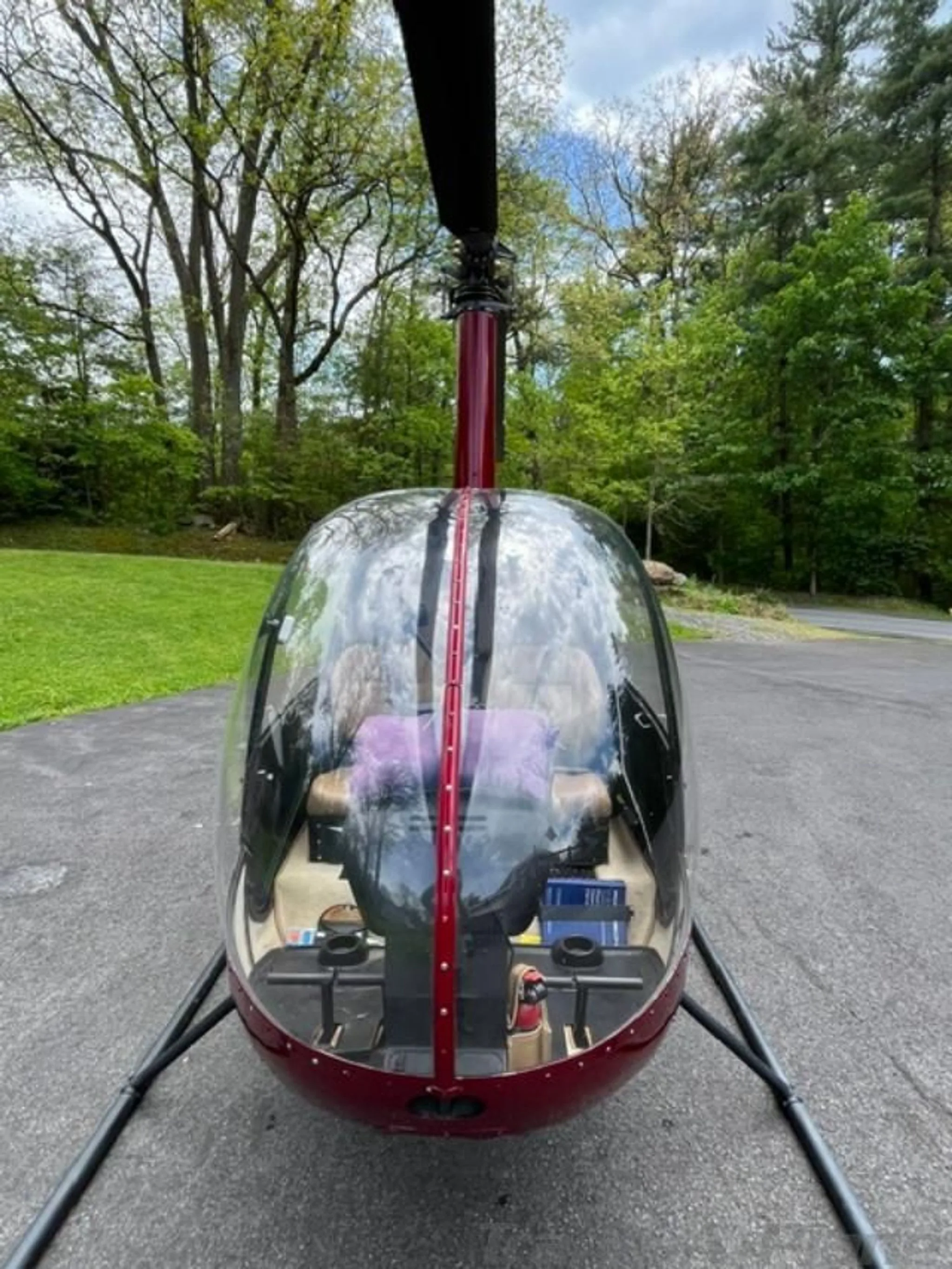 A front view of a red helicopter with a clear bubble cockpit, parked on a paved surface with trees in the background.
