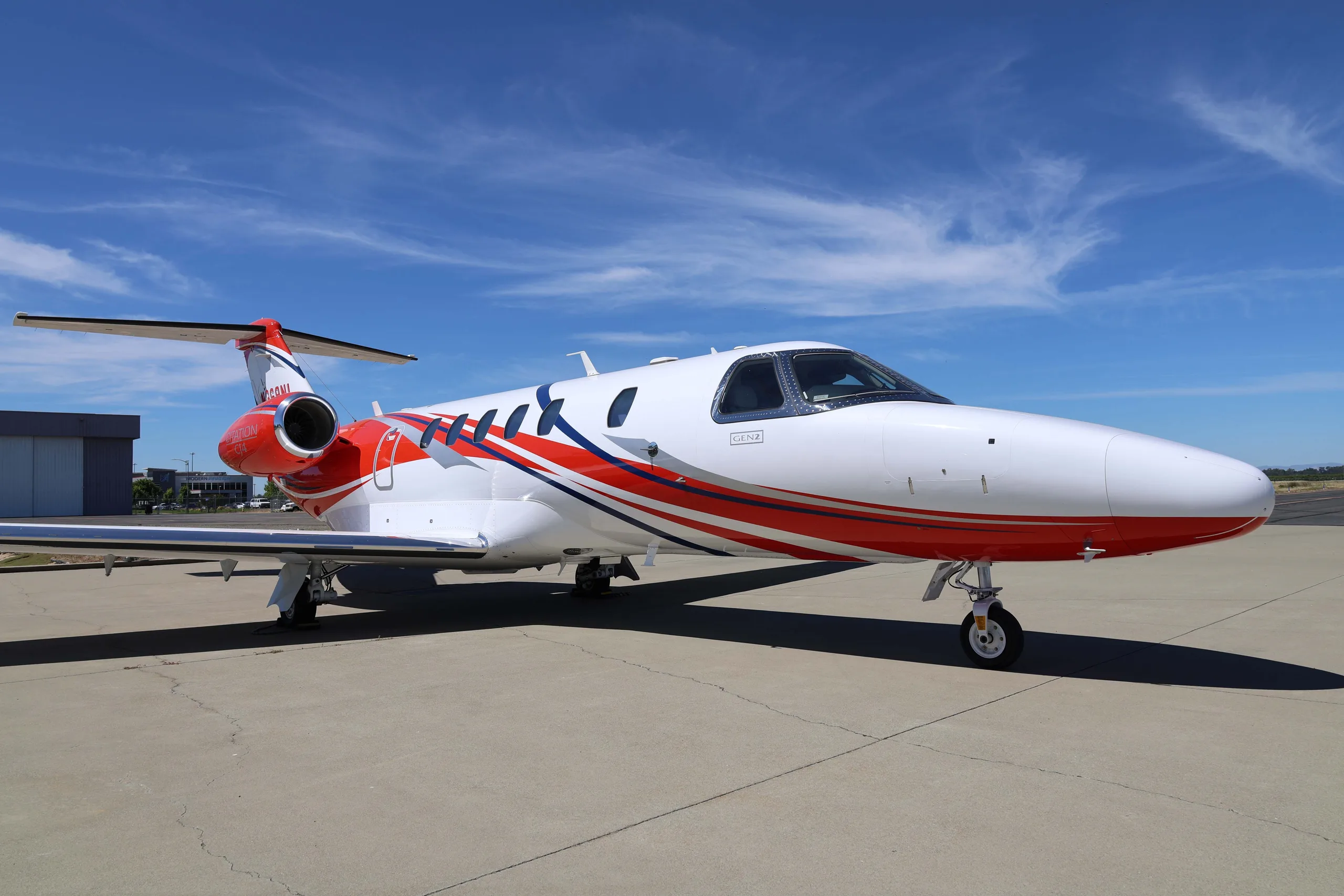 A white Cessna Citation CJ4 (N369NL) with red and blue stripes on the fuselage, parked on an airfield under a blue sky.