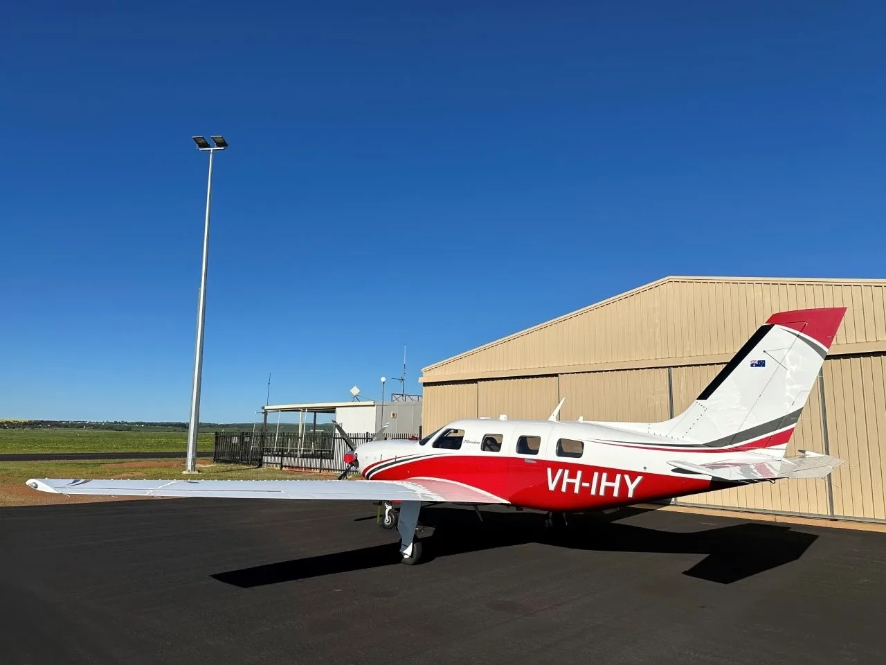 A white and red Piper Meridian aircraft with registration VH-IHY parked on a tarmac next to a hangar under a clear blue sky.