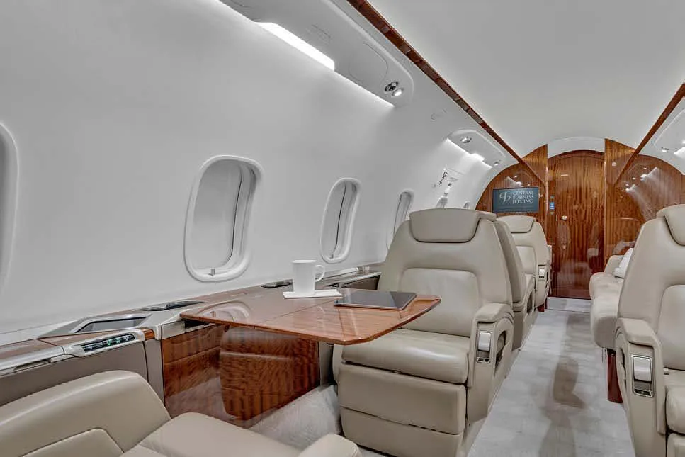 A view inside the aircraft cabin showing light beige leather seats, a polished wood table with a tablet and mug, and large windows.