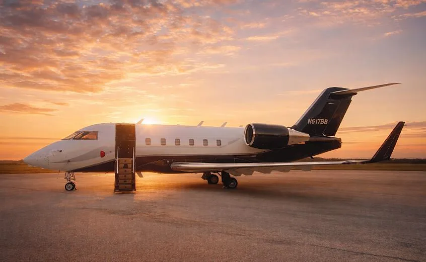 A white and dark blue Bombardier Challenger 350 business jet with registration N517BB is parked on a tarmac at sunset, with its main door open and stairs deployed.