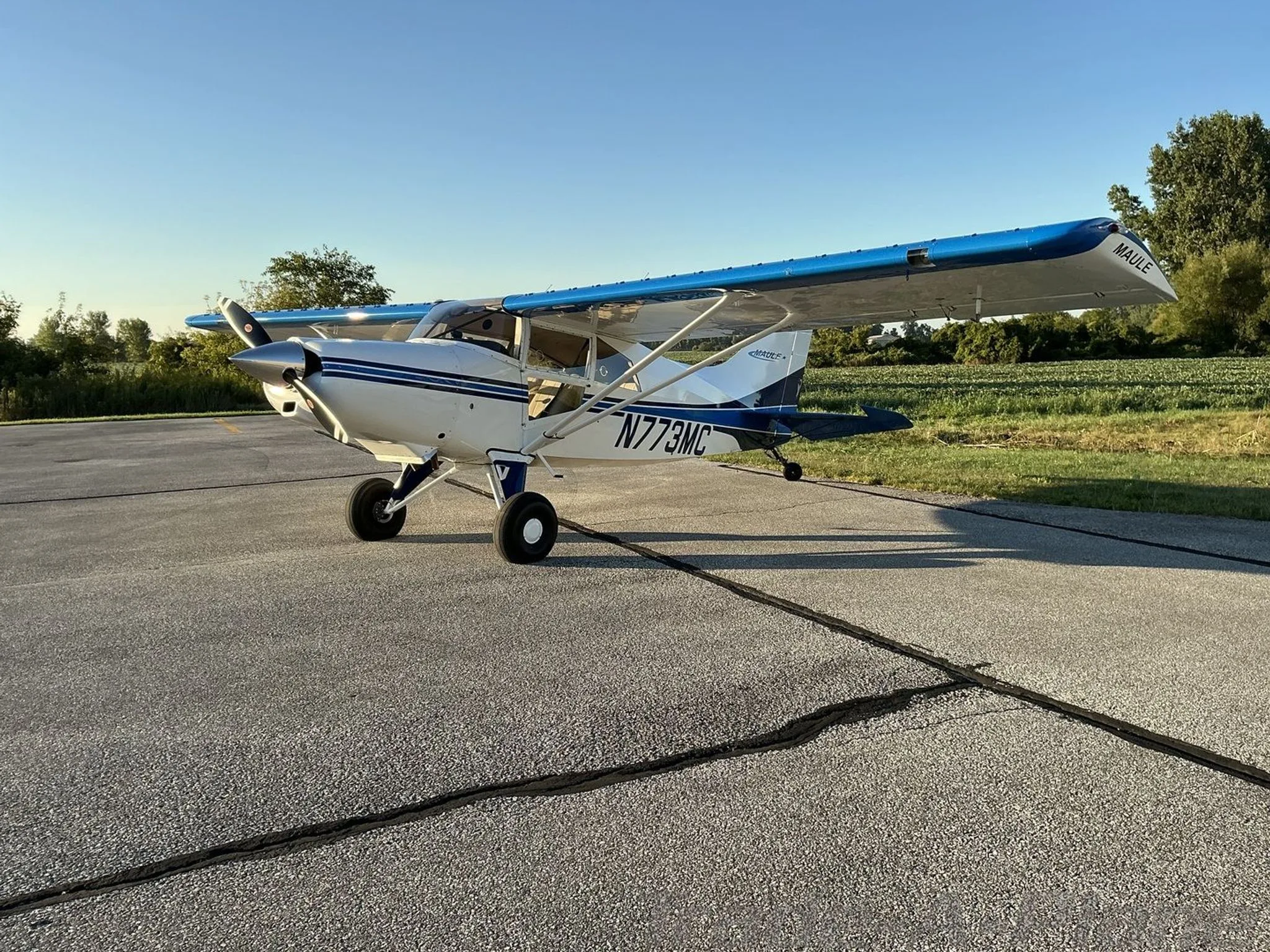 A white Maule aircraft with blue accents on the wings and tail, parked on an asphalt runway with fields in the background.