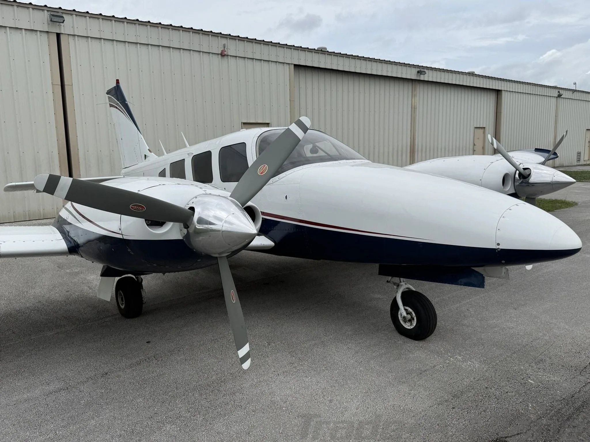 Front-left view of a white and blue twin-engine propeller aircraft with a red stripe, parked on an asphalt apron.