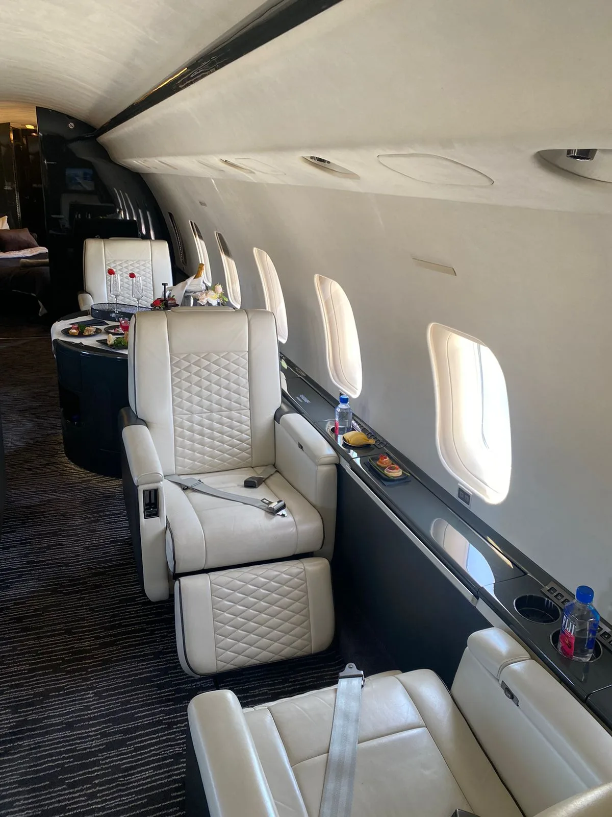 A luxurious private jet interior with white quilted leather seats and a table set with food and drinks.