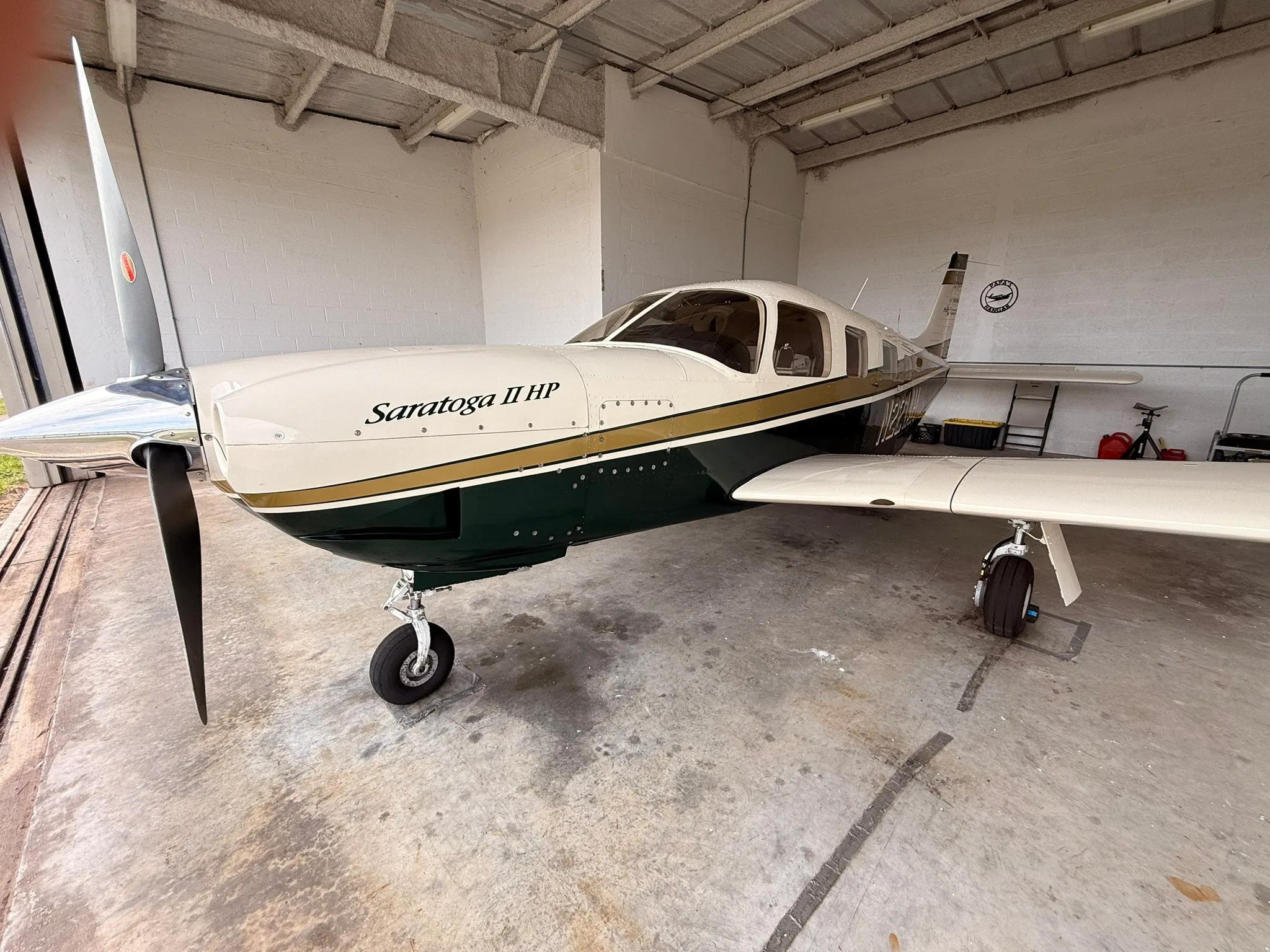 A Piper Saratoga II HP, N221AH, with a white, gold, and dark green paint scheme, parked inside a hangar.
