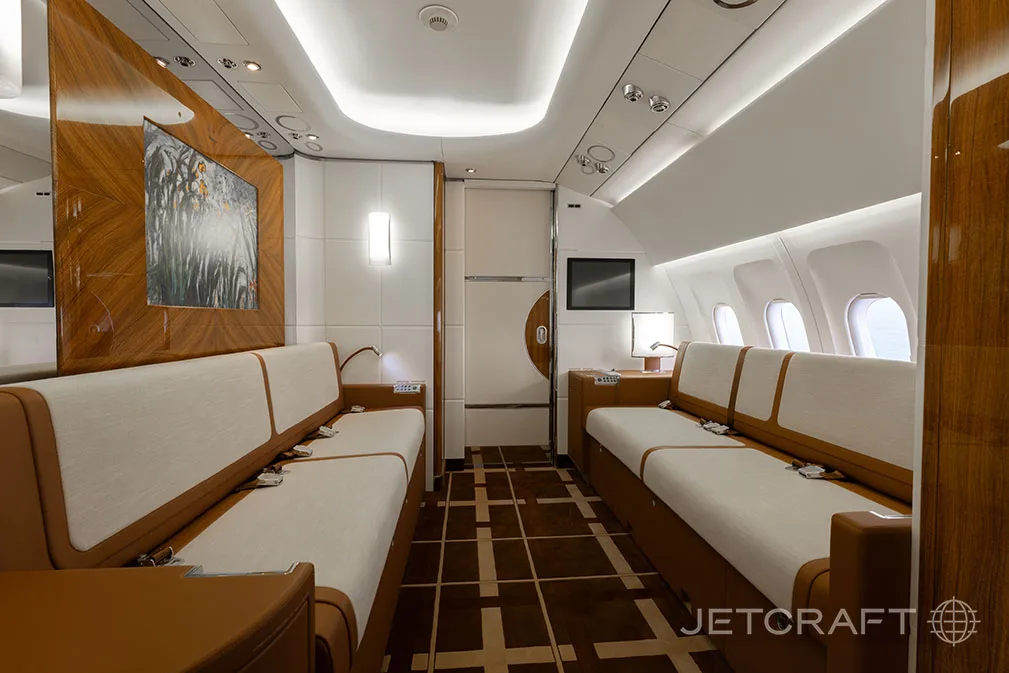 A spacious aircraft cabin featuring two long sofas facing each other, with wood paneling and a large wall-mounted artwork.