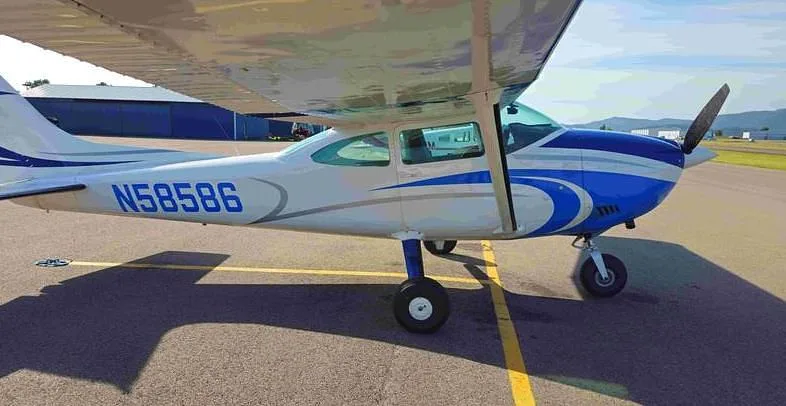 A side view of the white and blue Cessna N58586 aircraft parked on an asphalt tarmac with a hangar in the background.