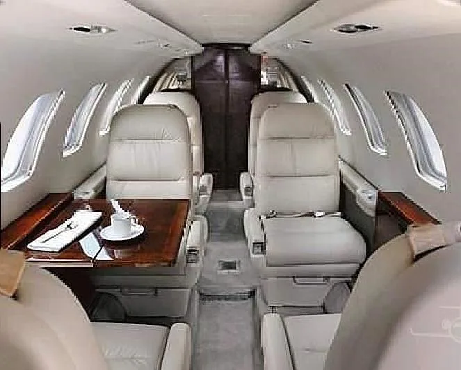 A wide view of the aircraft cabin interior showing multiple light-colored leather seats, a wooden table, and windows.