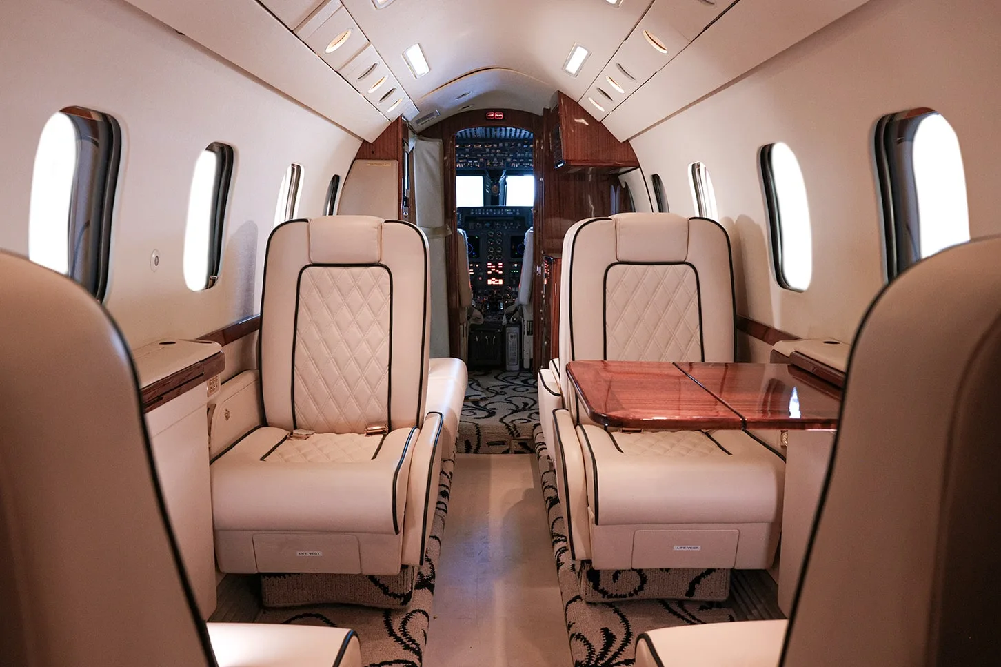 A view down the aisle of a private jet cabin, showing cream leather seats, a wooden table, and the cockpit beyond.