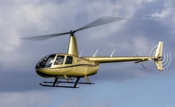 A gold Robinson R44 helicopter with a pilot visible, flying against a cloudy sky.