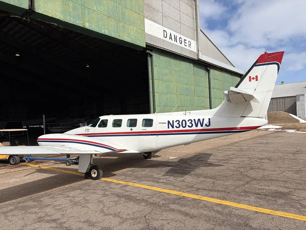 A side view of the N303WJ twin-engine aircraft with red and blue stripes, parked on an airfield next to a hangar.
