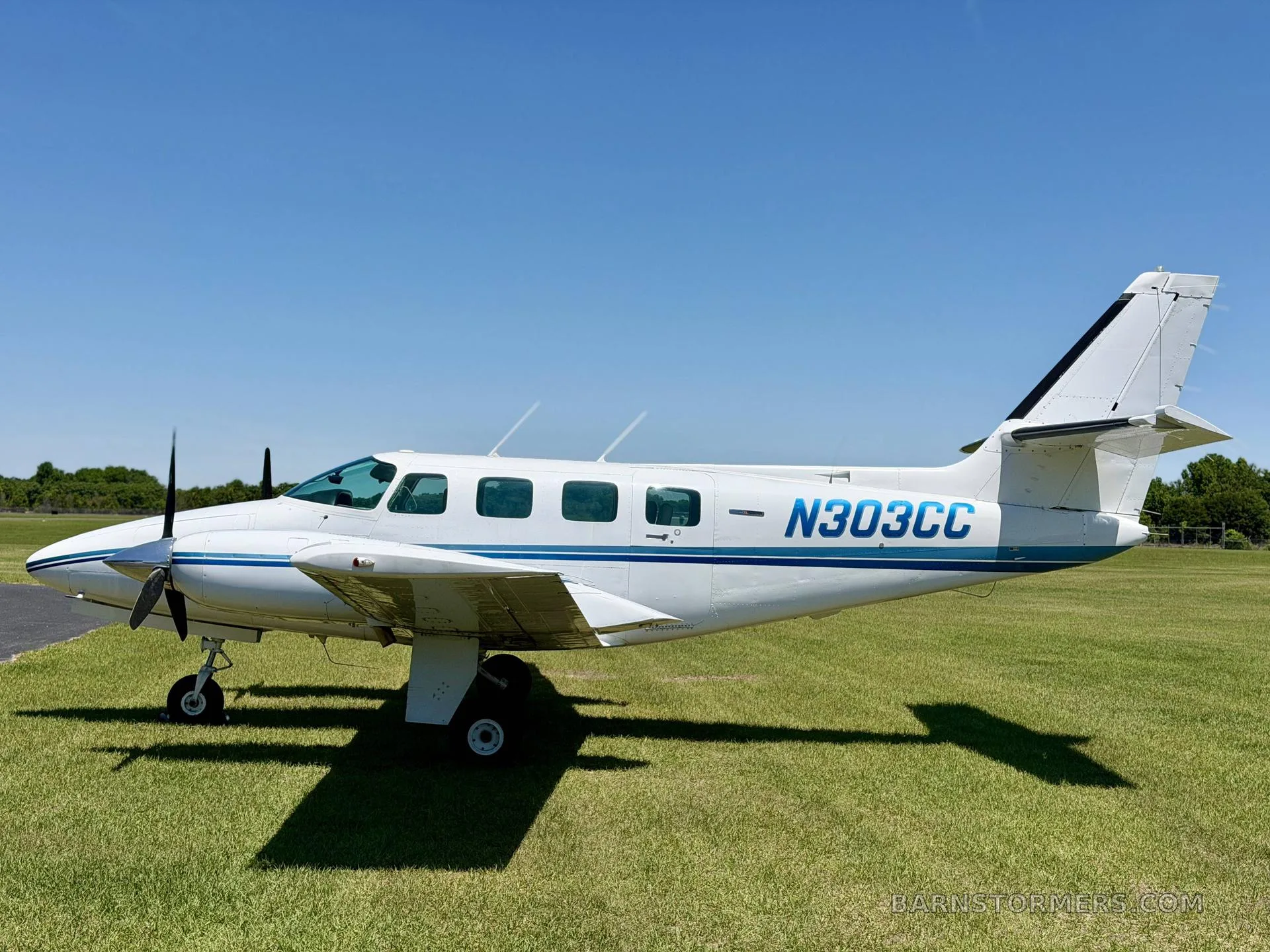 A white twin-engine propeller aircraft with blue stripes (N303CC) is parked on a grassy field, viewed from the side.