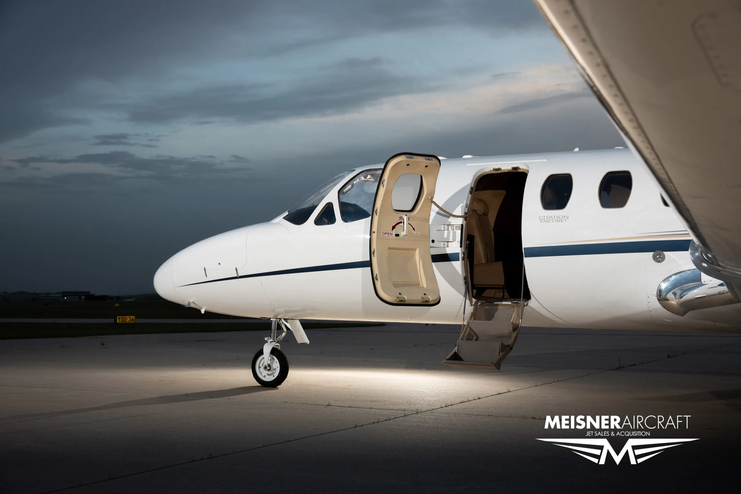 A close-up of the front of a white Cessna Citation jet with its main cabin door open and stairs extended, parked on an airfield.