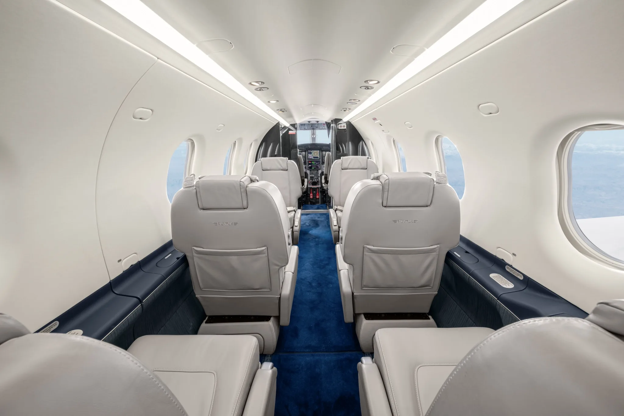 A view down the aircraft's aisle, showing multiple rows of light grey leather seats and a blue carpet.