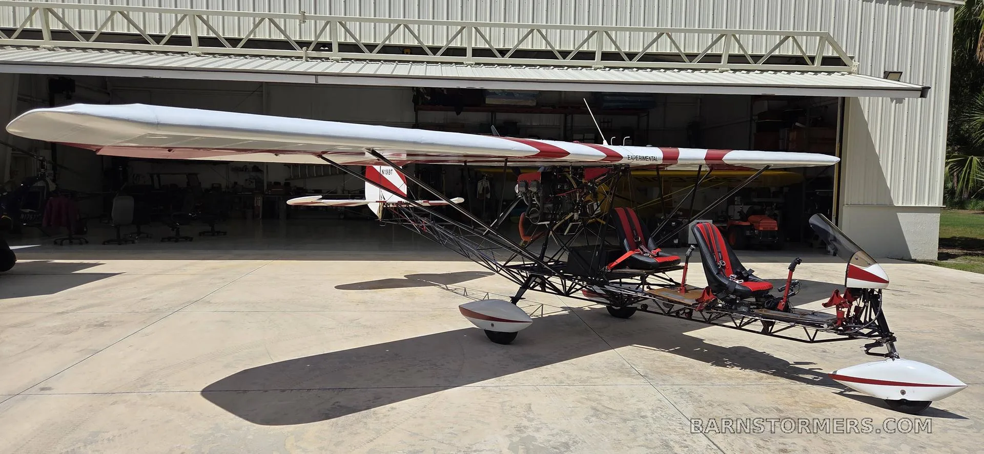 A red and white ultralight aircraft with an open frame is parked on concrete in front of an open hangar.