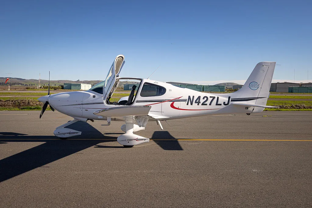 A white single-engine aircraft, N427LJ, with its gull-wing doors open, viewed from the side on an airfield.