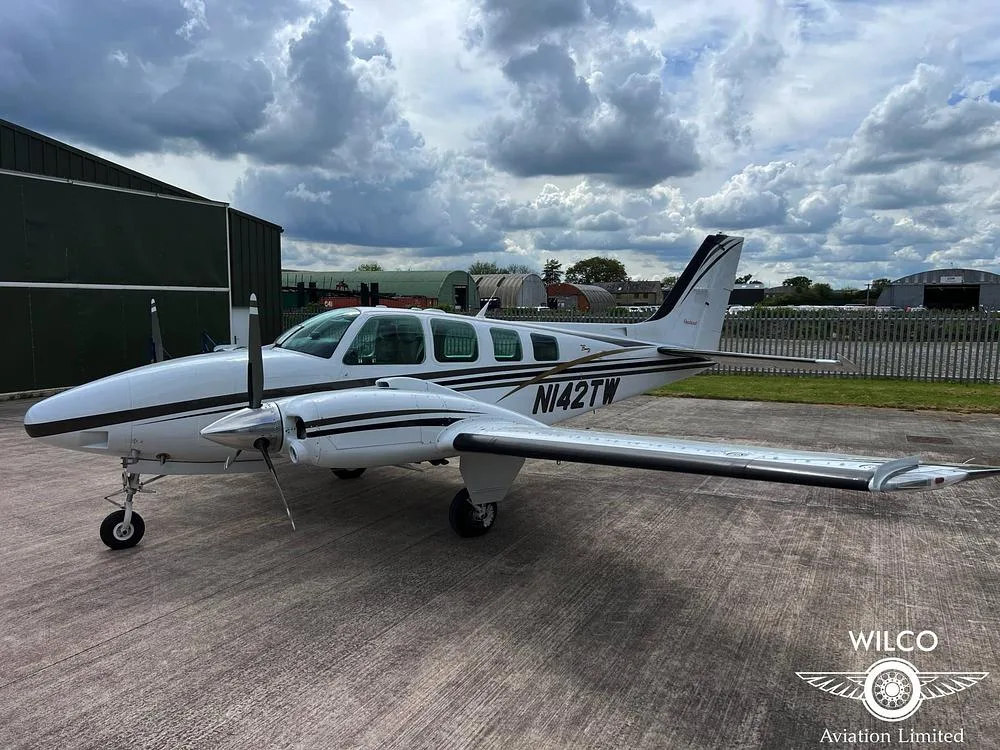A white Beechcraft Baron N142TW with black and gold stripes parked on a tarmac with a fence in the background.