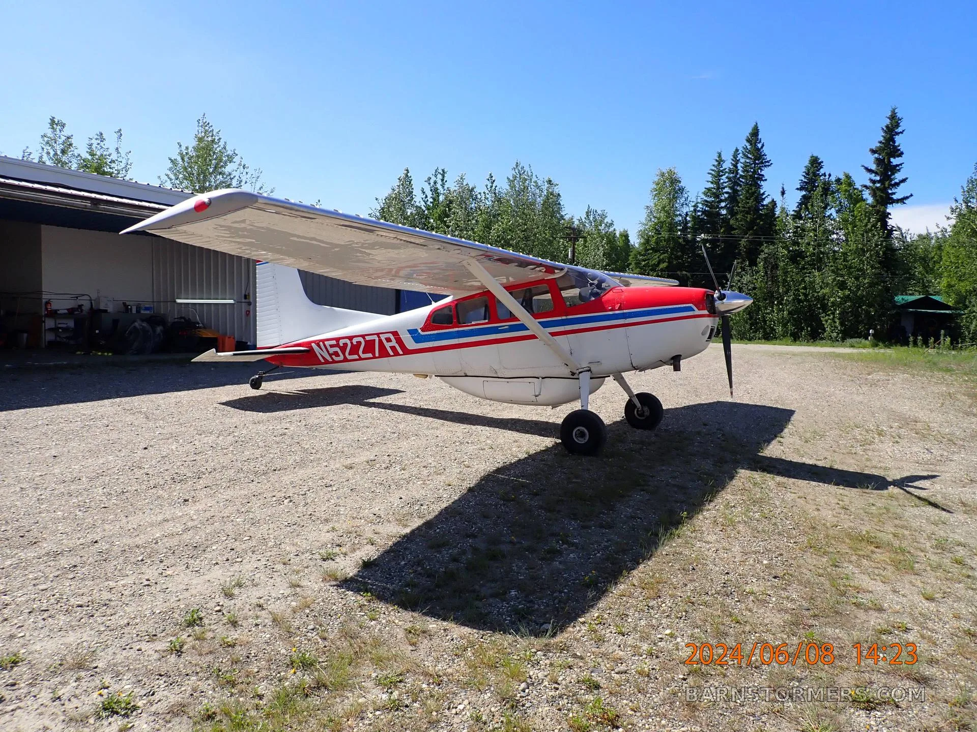 A side view of a red, white, and blue Cessna 180 aircraft with registration N5227R parked on a gravel lot next to a hangar.