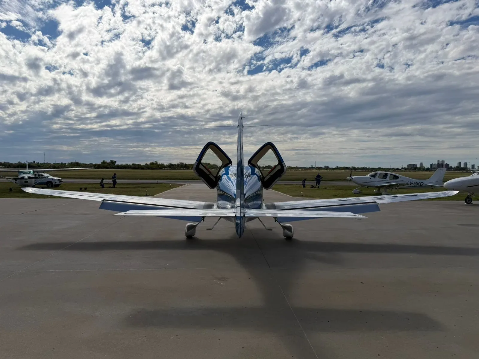 A blue and silver Cirrus SR22 aircraft with its gull-wing doors open, viewed from the rear on an airfield.