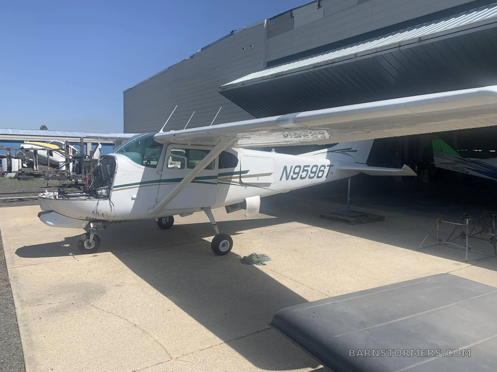 A white Cessna 182 aircraft with green and gold stripes, registration N9596T, parked outside a hangar with its engine cowling removed.