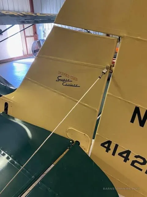 A close-up view of the tail of a beige and dark green Piper Cub Super Cruiser aircraft, showing its model name and registration number.