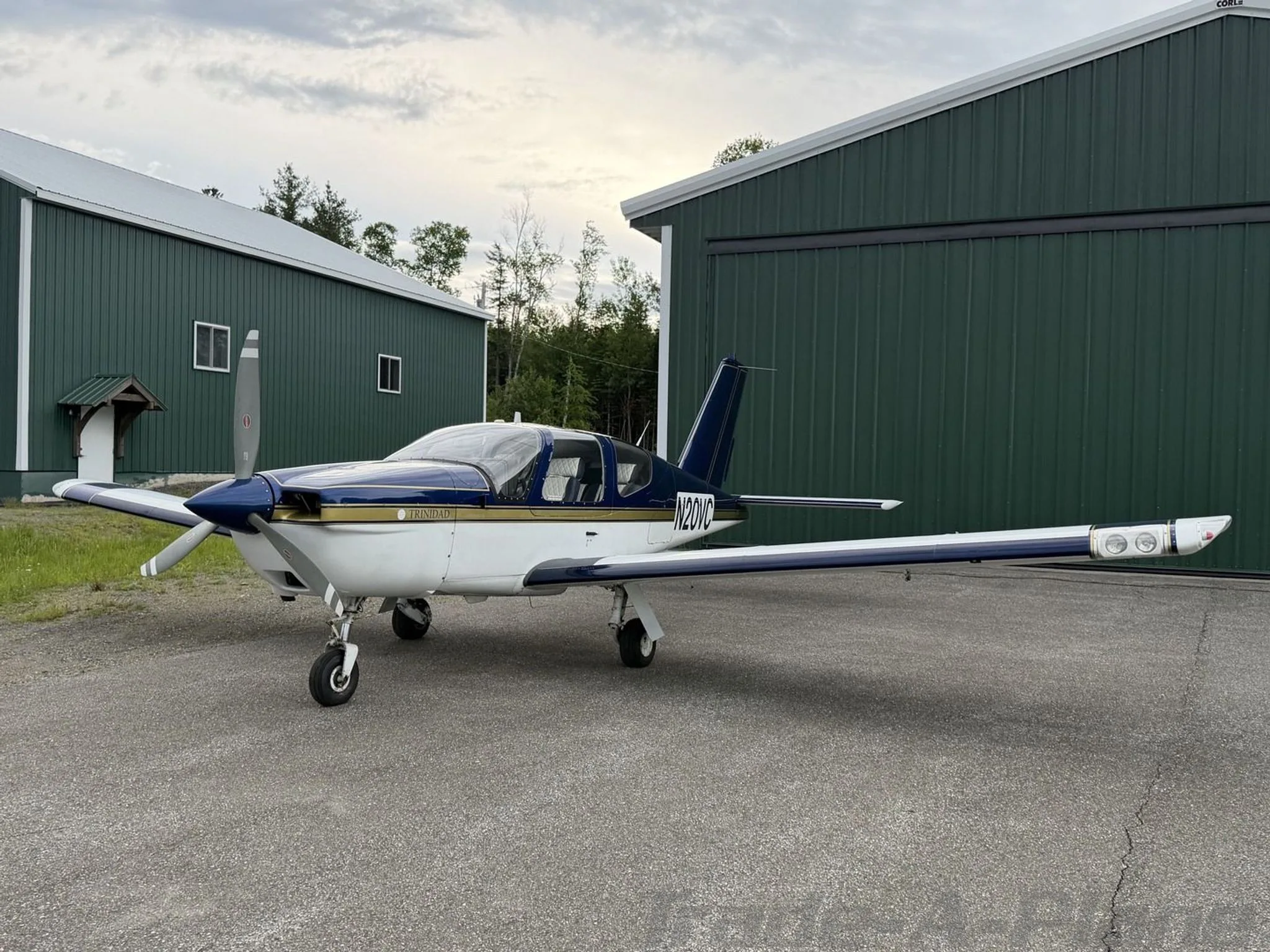A white and blue Socata Trinidad TB20 aircraft with gold trim, tail number N20VC, parked on an asphalt apron next to green hangars.