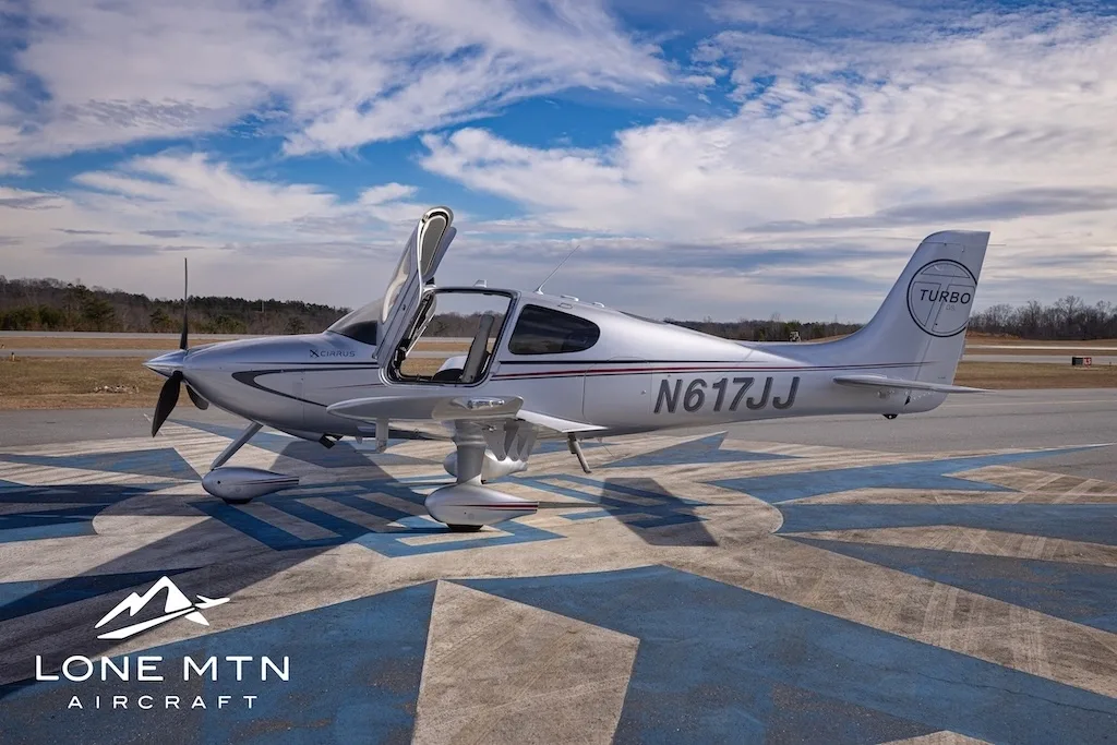 A silver Cirrus SR22T aircraft, N617JJ, with its doors open, parked on an airfield from a side view.