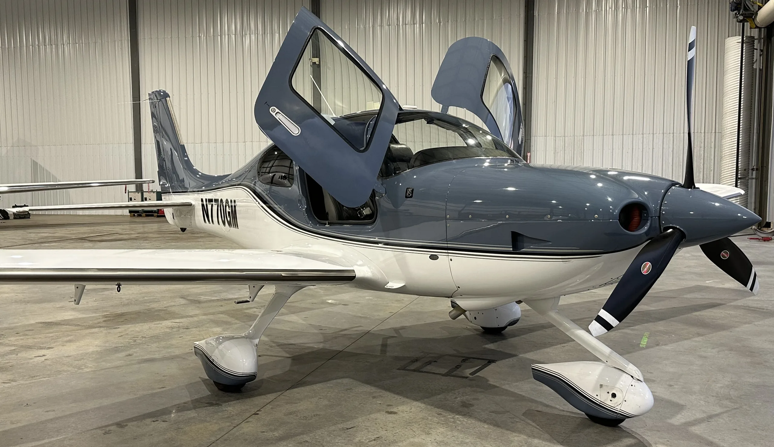 A grey and white Cirrus SR22 aircraft with registration N770GM, parked in a hangar with its gull-wing doors open.