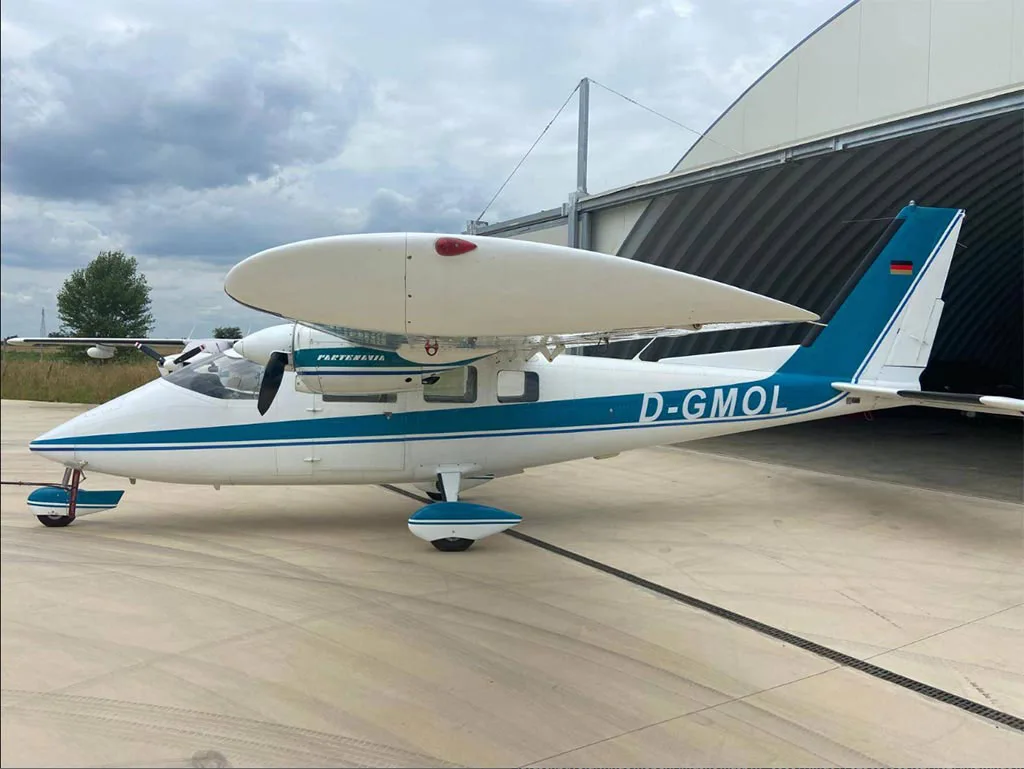 A white and blue Partenavia P.68 aircraft with registration D-GMOL parked on an airfield next to a hangar.