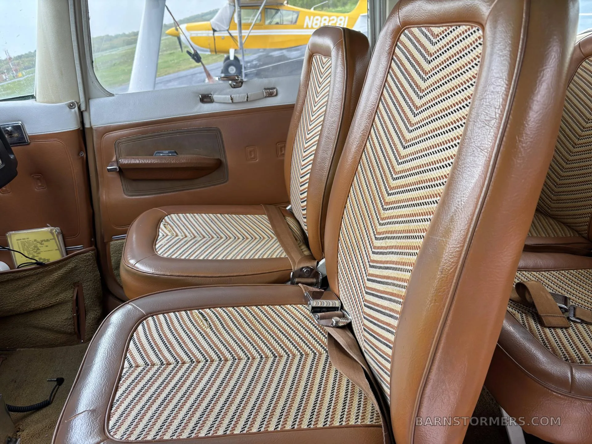 The brown leather and patterned fabric seats of a small aircraft interior, with a view of another aircraft outside.